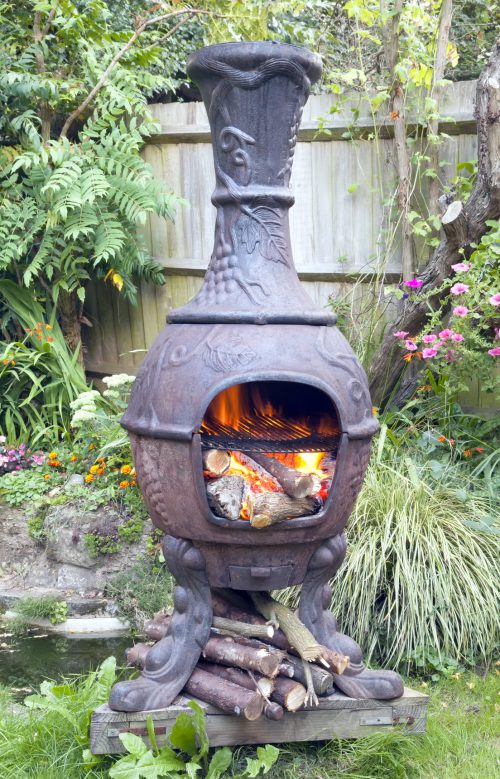 Rustic cast iron Mexican chiminea style wood burning stove with flames, in summer cottage flower garden